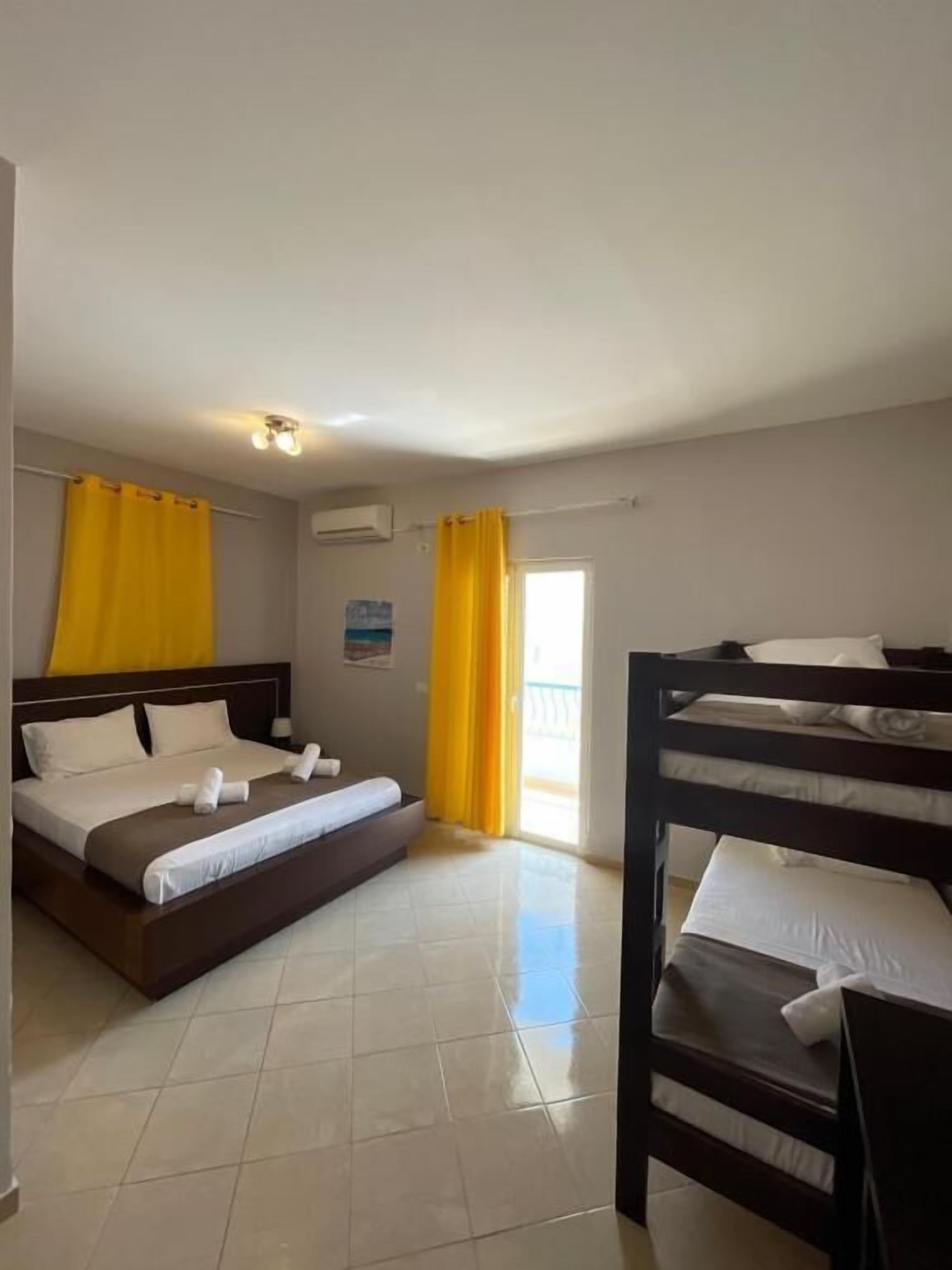 Quarto de hotel com uma cama de solteiro, uma cama beliche e uma janela com cortinas amarelas.