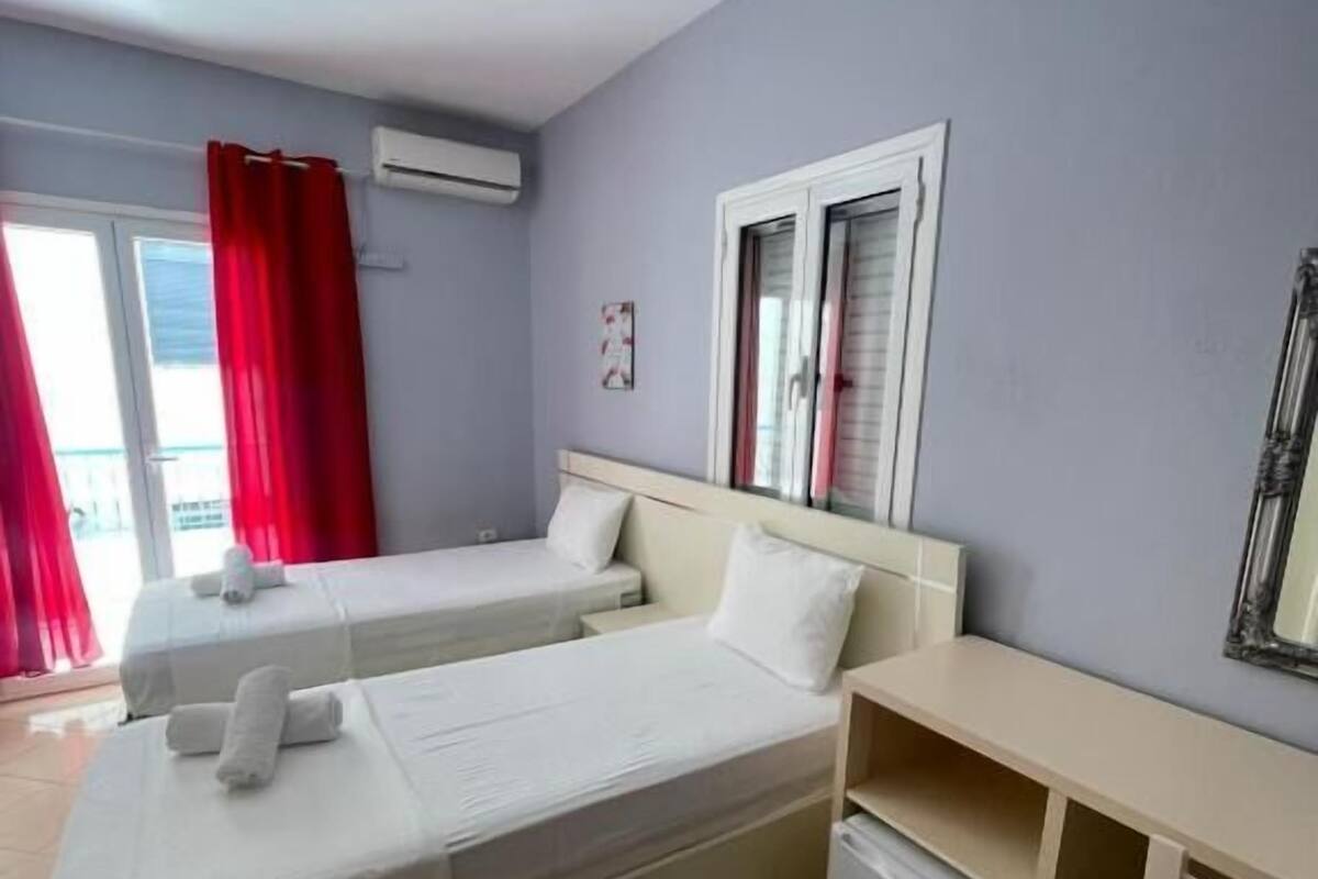 A hotel room with two beds, a desk, a chair, a ceiling fan, and a window with red curtains.