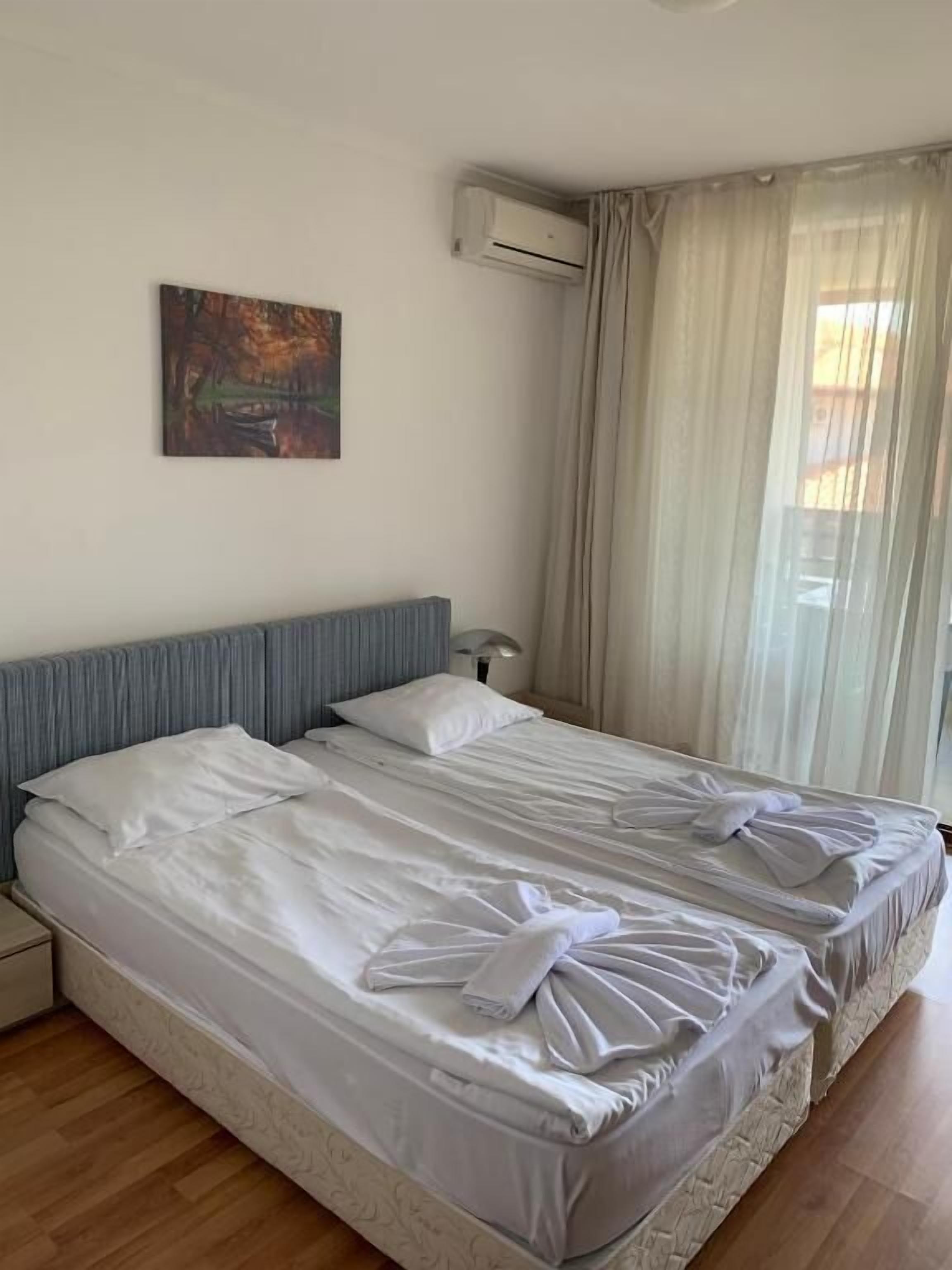 A double bed with white linens and towels, a headboard, an air conditioning unit, a painting on the wall, and a window with curtains.