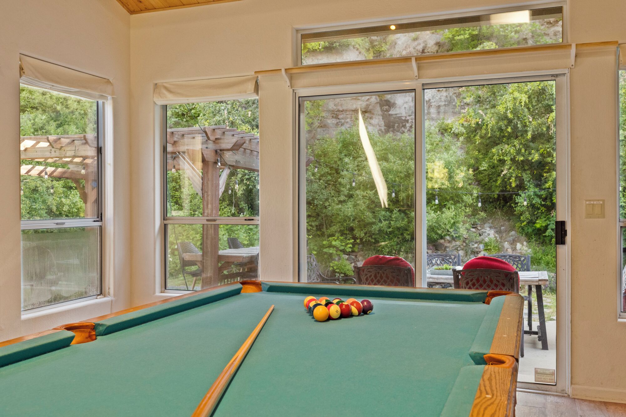 Un tavolo da biliardo con le palle, una pergola in legno all'esterno e vista sul verde attraverso ampie finestre.