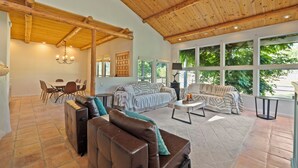 A spacious living area with a high wooden ceiling, a dining table with chairs, a sofa, and a large window with a view of greenery.