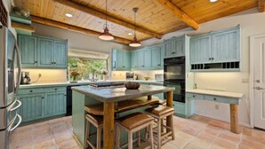A kitchen with teal cabinets, a large island with stools, a wooden ceiling, and a view of greenery outside.
