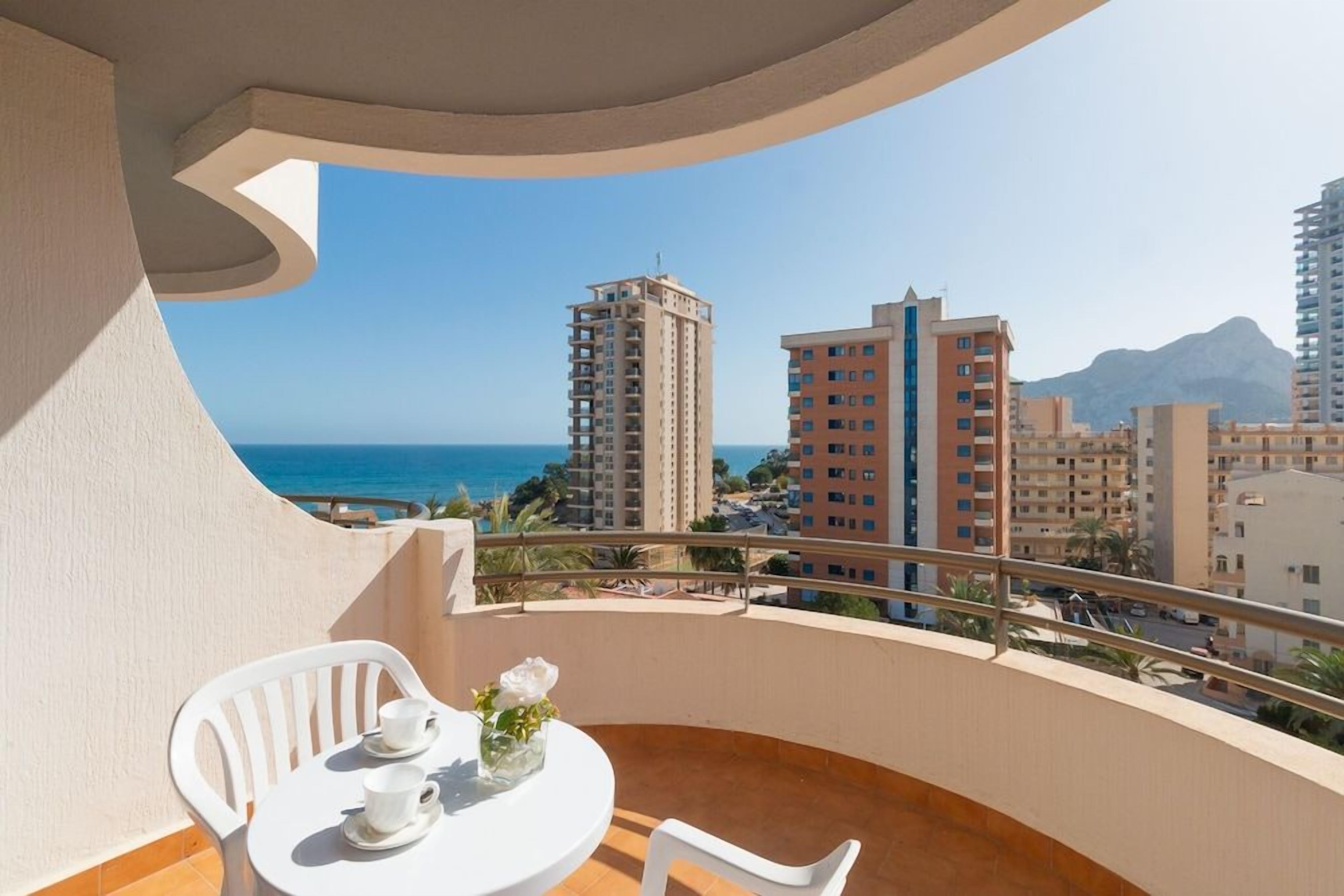 Un balcón con mesa y sillas, con vistas a un paisaje urbano que incluye edificios y una masa de agua.