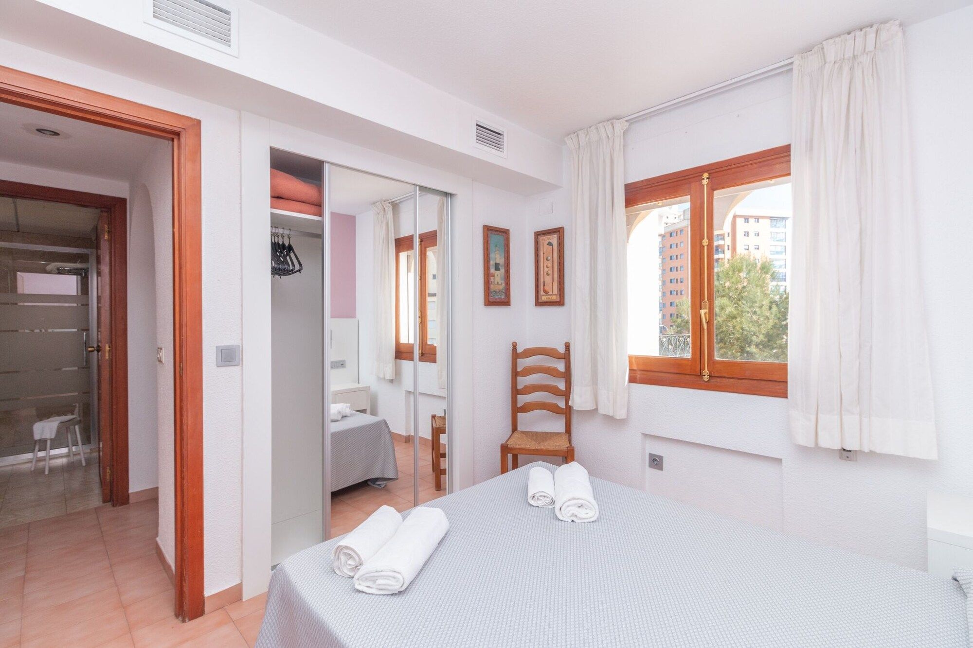 Un dormitorio con cama, una silla, ventana con cortinas, armario y un espejo.