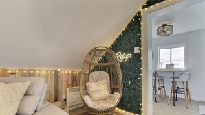 Ein gemütliches Wohnzimmer mit Korbsessel, Sofa und einer Wand, die mit Grünpflanzen und Lichterketten dekoriert ist.