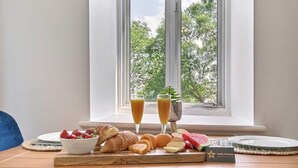 Desayuno con cruasanes, fresas, sandía, queso y dos vasos de jugo de naranja sobre una mesa de madera junto a una ventana.