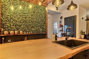 Una cocina con encimera de madera, un lavamanos negro y una pared cubierta de follaje verde. Hay luces colgantes y un refrigerador al fondo.