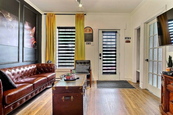 Un salón con un sofá de cuero, una mesa de centro de madera, un sillón estampado, una maleta antigua y una puerta con ventanal.
