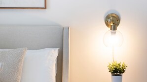 Un lit bien fait, avec du linge de lit blanc, une table de chevet ornée d’une plante en pot et de quelques livres, et une lampe fixée au mur.