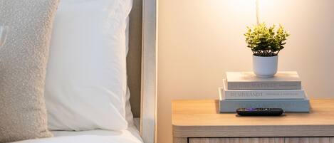 A neatly made bed with white linens, a bedside table with a potted plant and books, and a wall-mounted lamp.