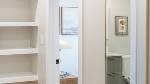 Un couloir aux murs blancs, doté d’un plancher en bois et agrémenté d’un miroir à cadre doré. On aperçoit des étagères sur le côté gauche, et une porte donne accès à une autre pièce.