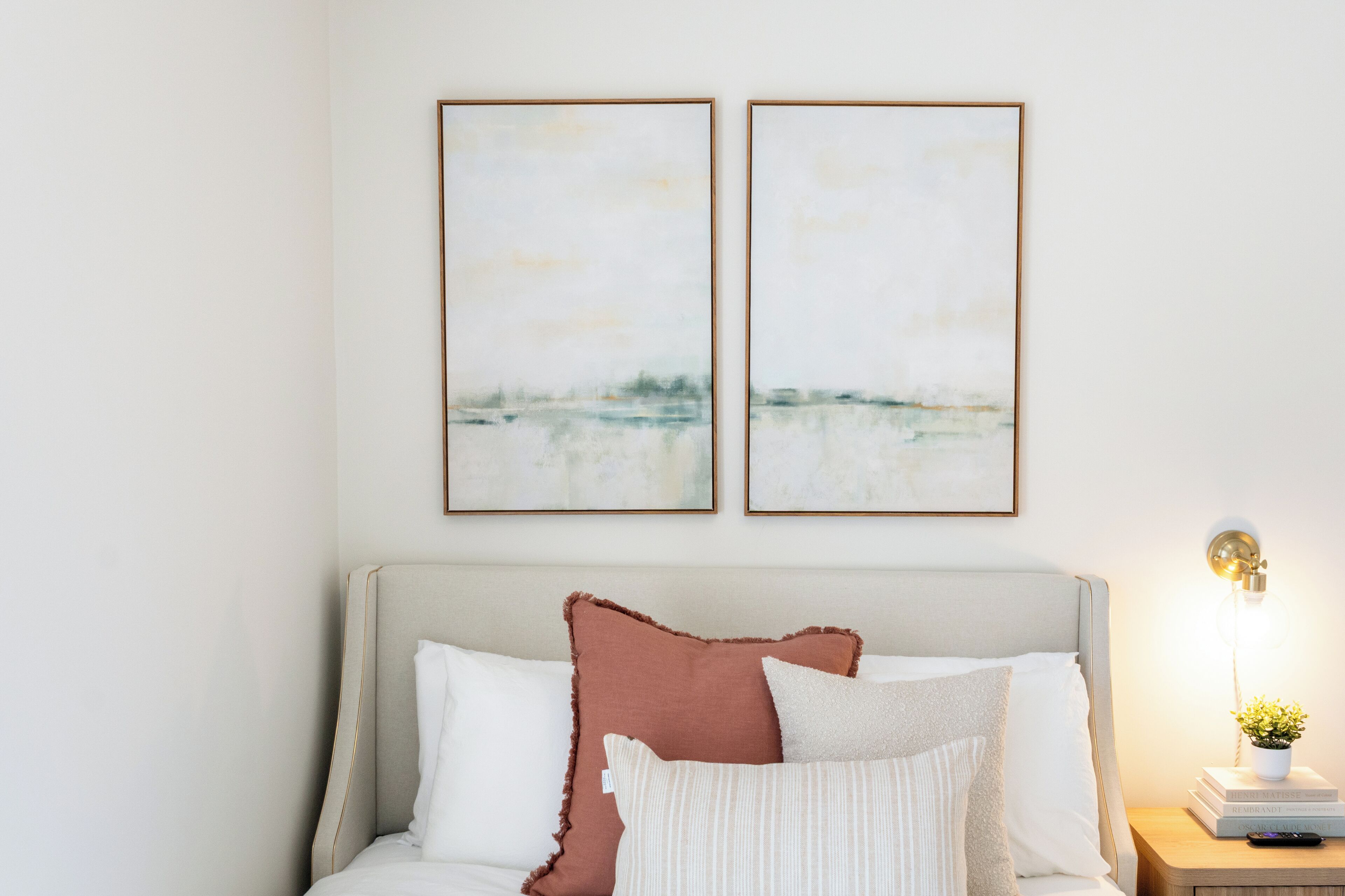 Ein ordentlich eingerichtetes Schlafzimmer mit einem Bett, Kissen, einem Nachttisch mit Lampe und zwei gerahmten Kunstwerken an der Wand.
