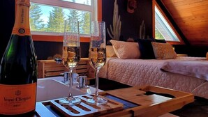 Une pièce avec un plafond en bois, un lit recouvert d’une courtepointe, deux coupes de champagne et une bouteille étiquetée « Veuve Clicquot ».