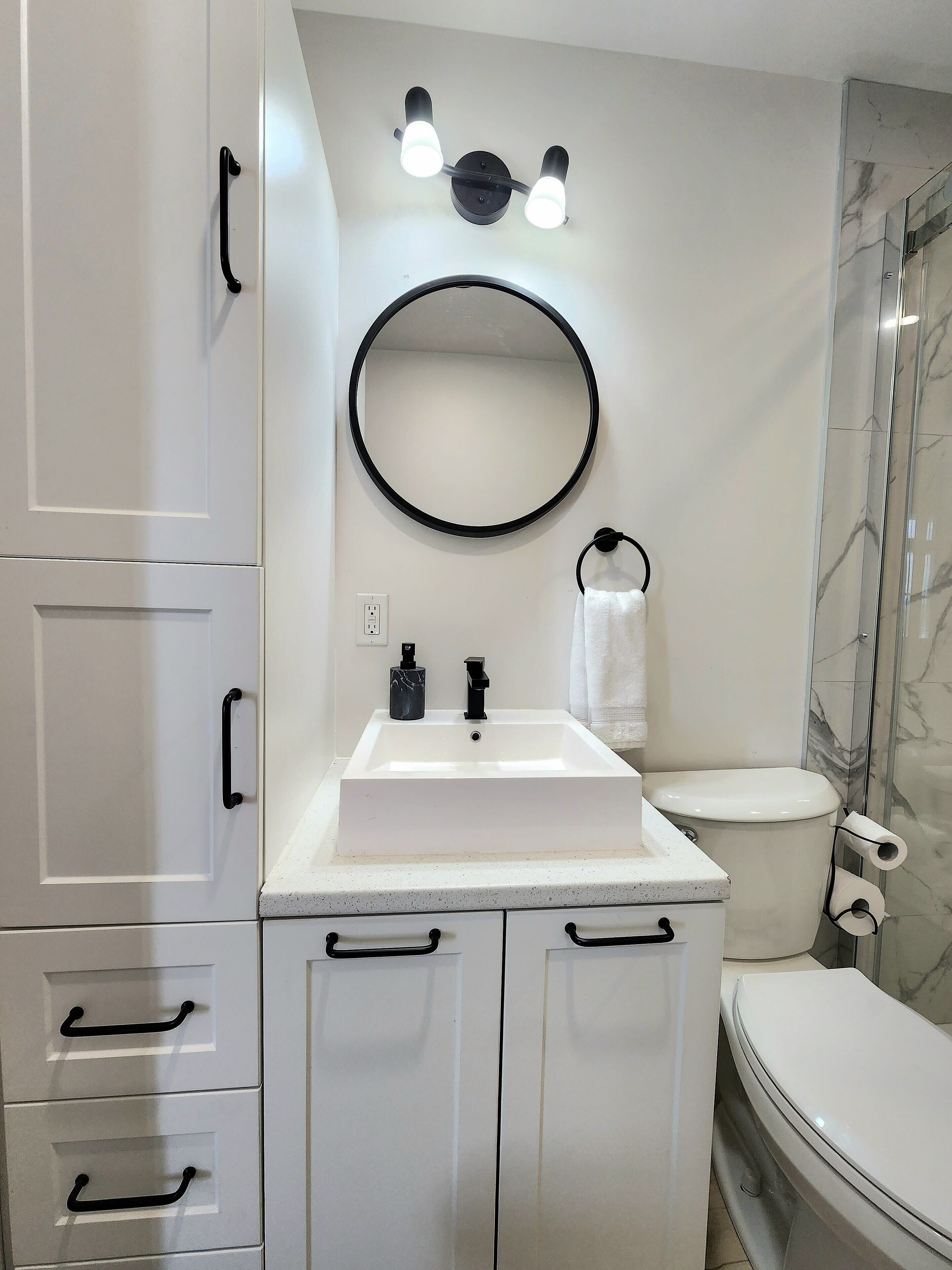 Een moderne badkamer met een wit wastafelmeubel, een zwarte kraan en een ronde spiegel erboven. Aan de rechterkant staat een toilet met een douchecabine met marmeren wanden.