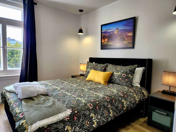 Un lit bien fait, recouvert d’une couvre-lit à motifs floraux, avec un oreiller jaune et une serviette blanche. Un tableau représentant un paysage urbain est accroché au mur, au-dessus du lit.