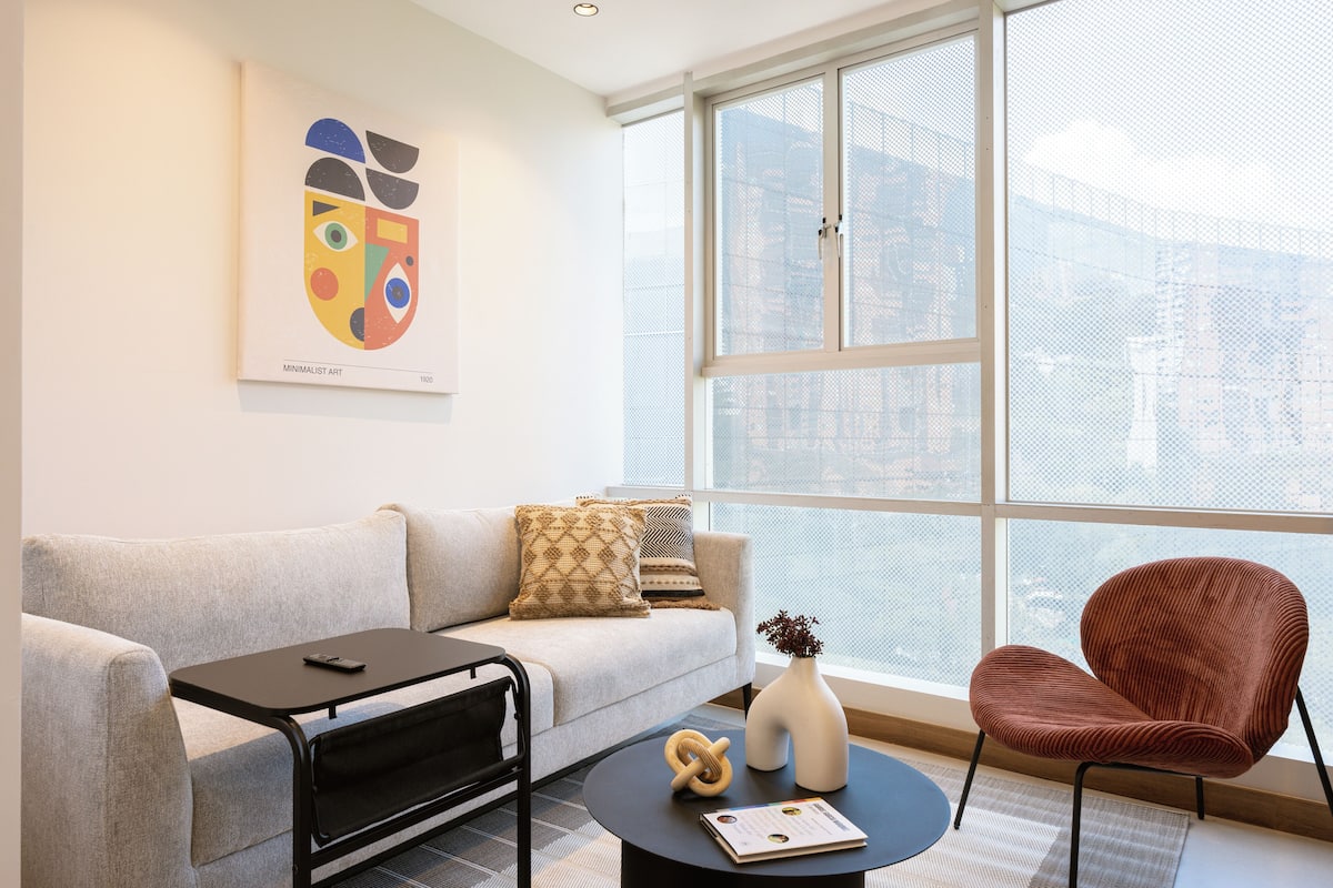 A modern living room with a sofa, a coffee table, a red chair, and a large window offering a city view.