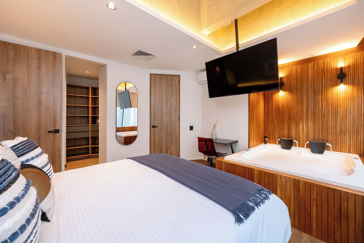 A modern hotel room with a large bed, a wooden wardrobe, a TV mounted on the wall, and a bathroom with a large bathtub and two sinks.