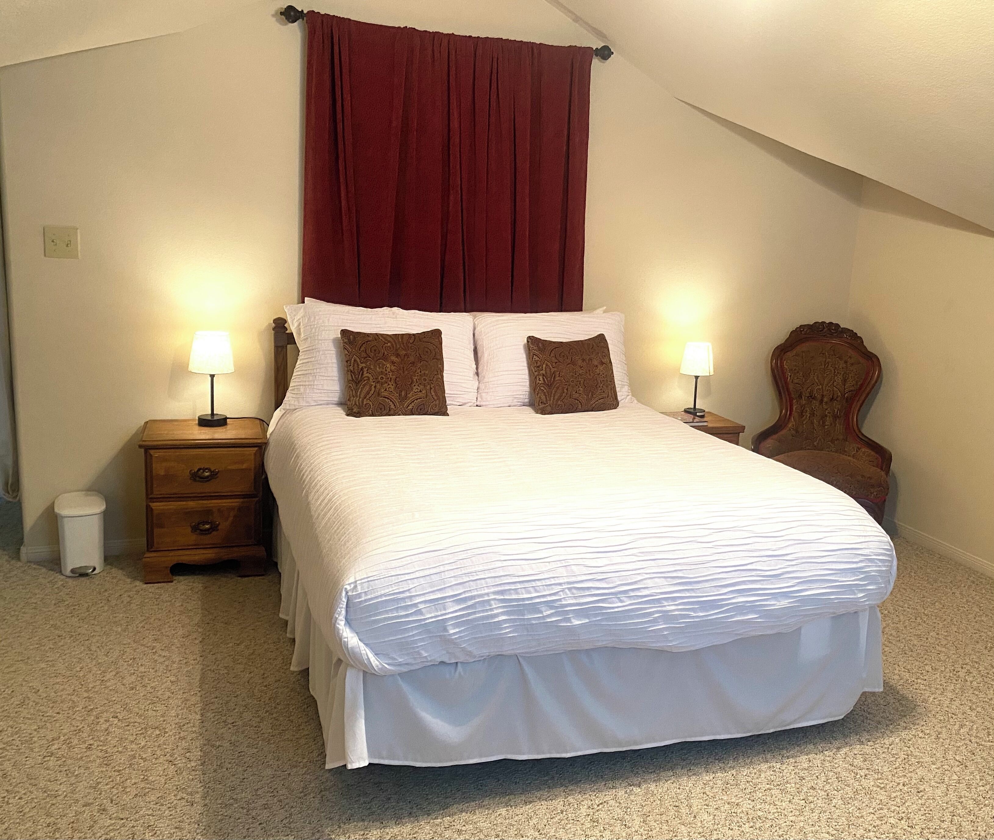 Una cama bien hecha con sábanas blancas y dos almohadones decorativos, una mesita de noche de madera con una lámpara, y una silla con estructura de madera.