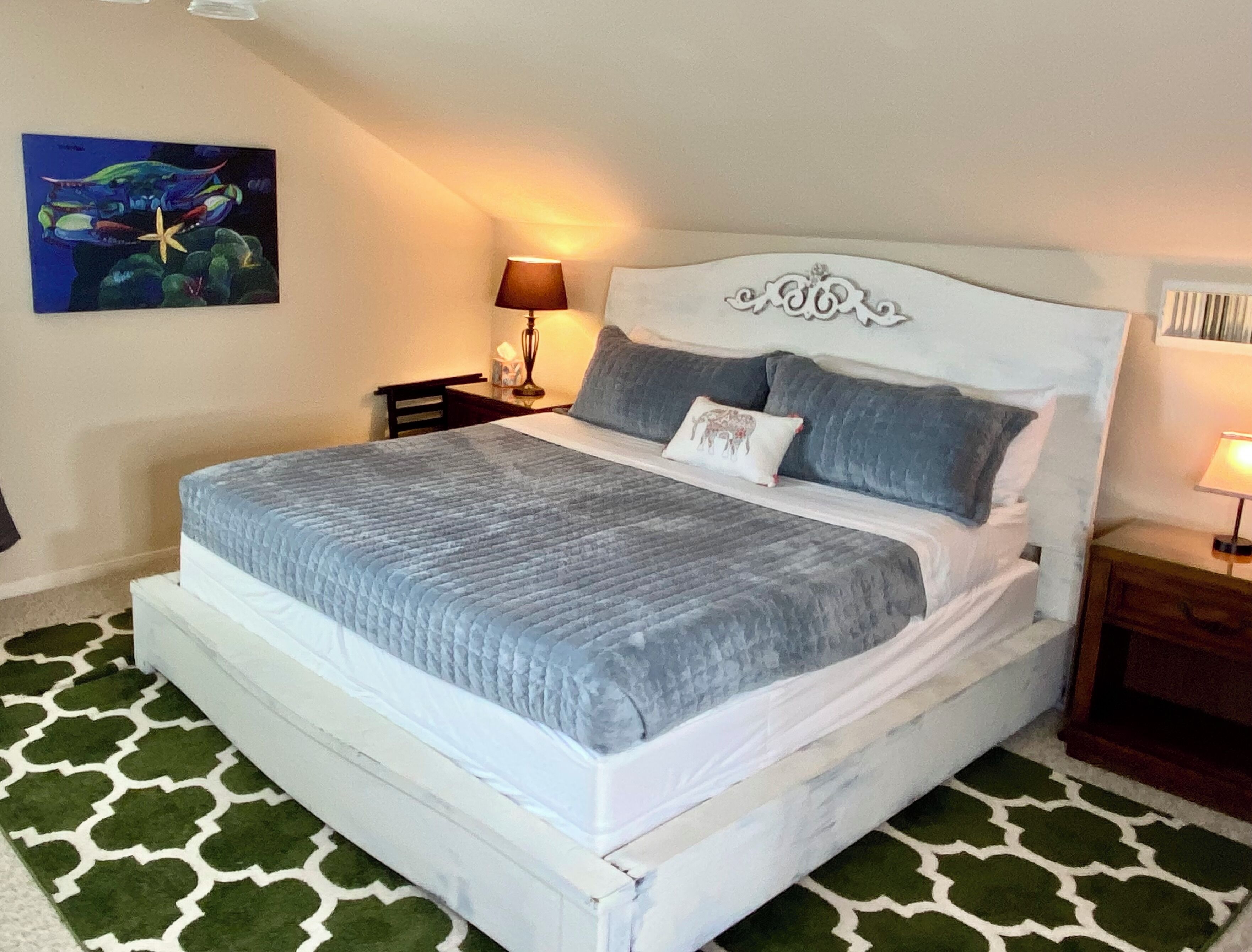 A bed with a white headboard, grey bedding, and a green patterned rug.