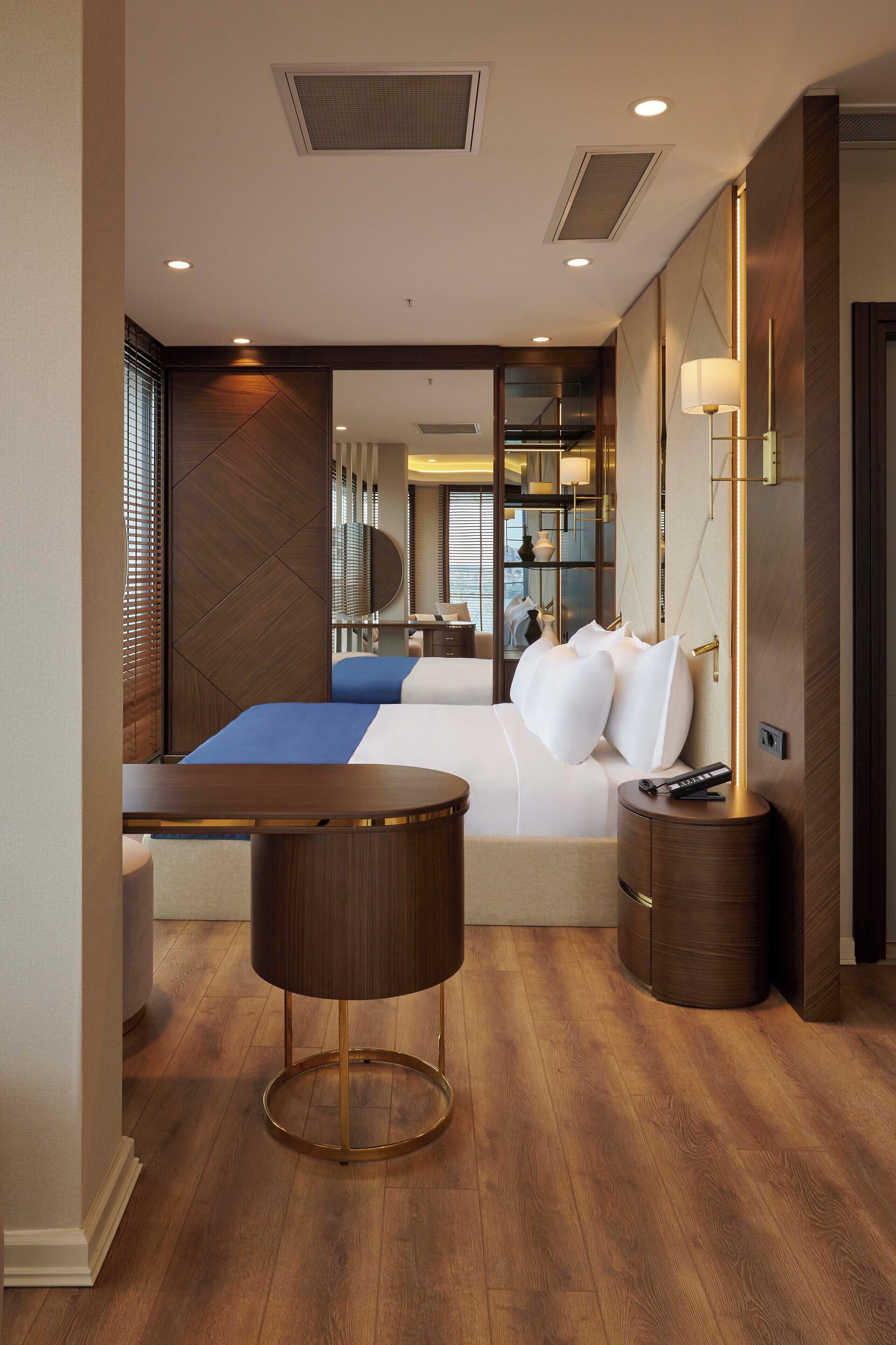 A modern hotel room with a large bed, a round wooden side table, a small round table, and a mirror on the wall.