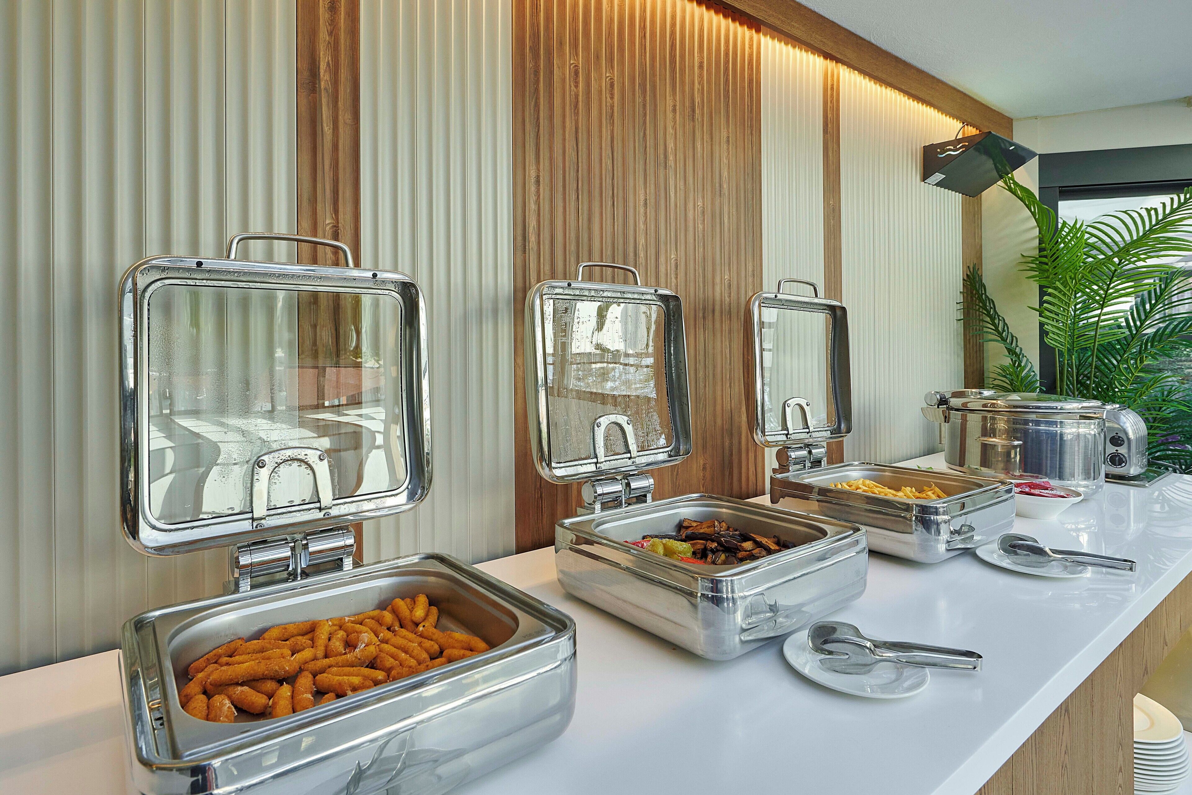 Disposición de un bufé con diversos alimentos en recipientes metálicos, una encimera blanca y un panel de pared de madera.