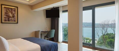 A hotel room with a bed, a desk, a chair, a TV, and a large window with a view of the sea.