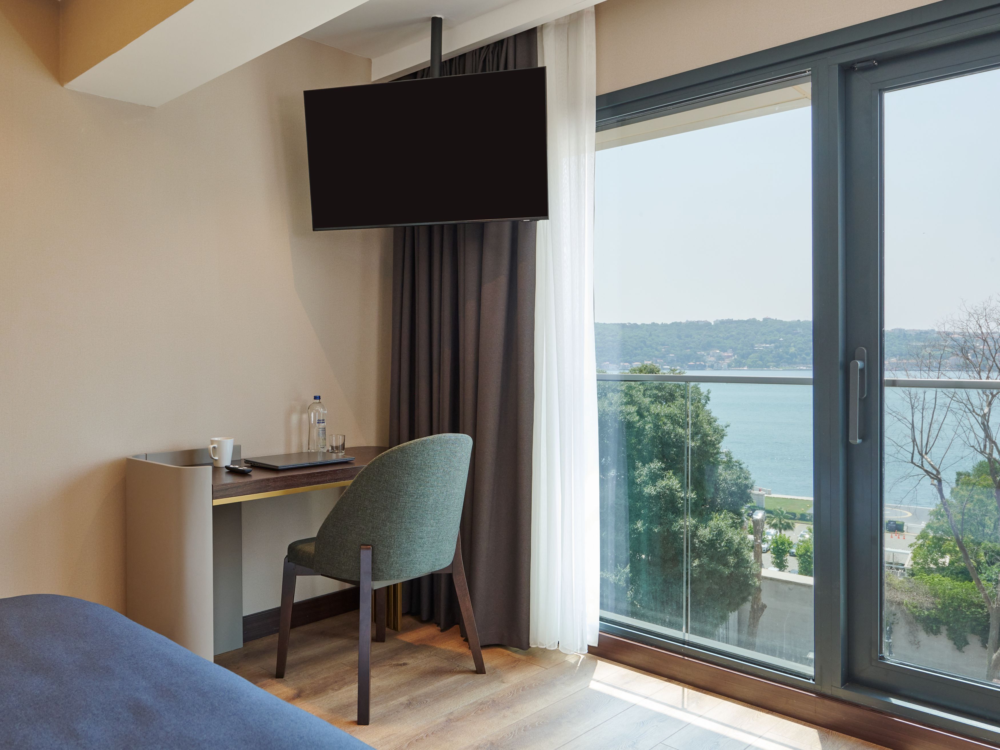 A hotel room with a bed, a desk with a chair, a flat-screen TV mounted on the wall, and a large window offering a view of a body of water and greenery.