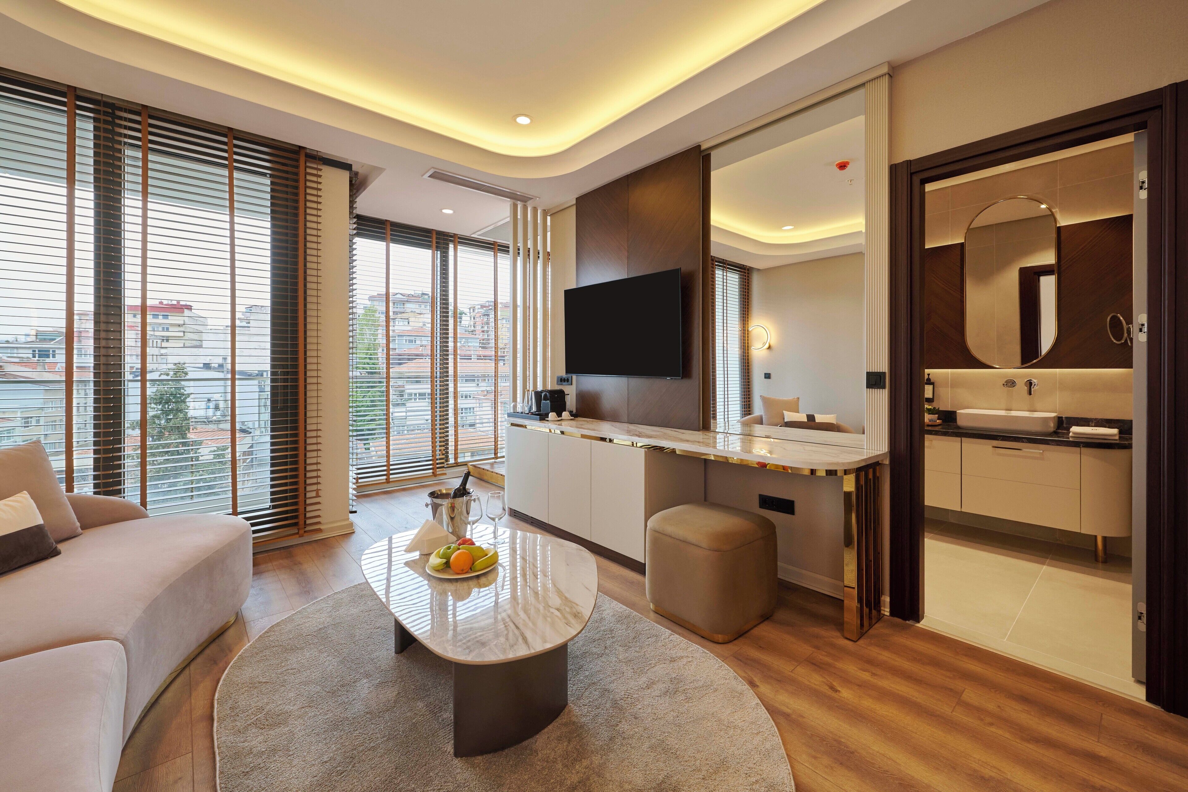 A modern hotel room with a large window, a flat-screen TV, a small kitchenette, and a bathroom with a mirror and sink.