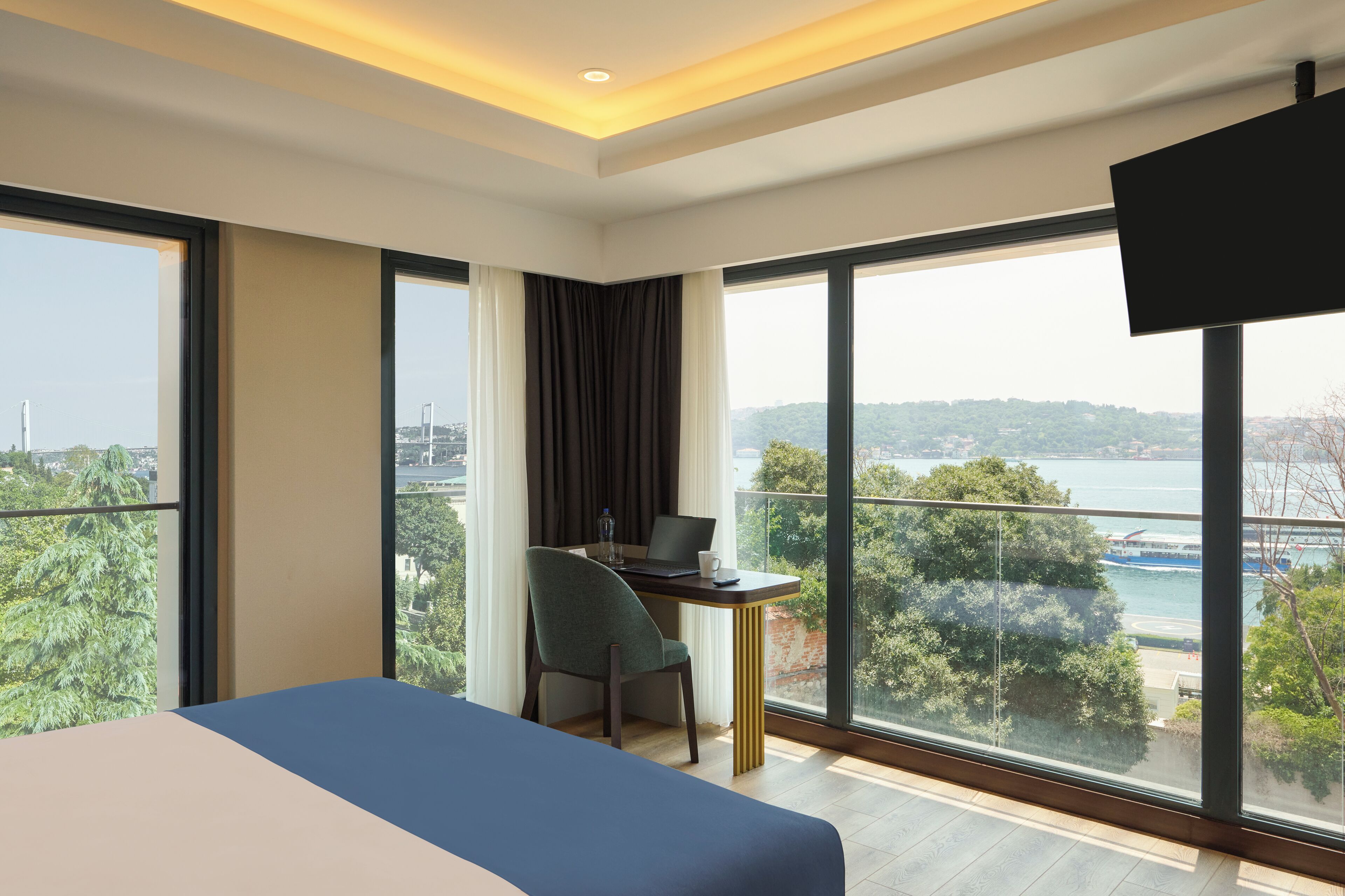 A hotel room with a large bed, a desk with a laptop, and a TV. The room has large windows with a view of a body of water and greenery.
