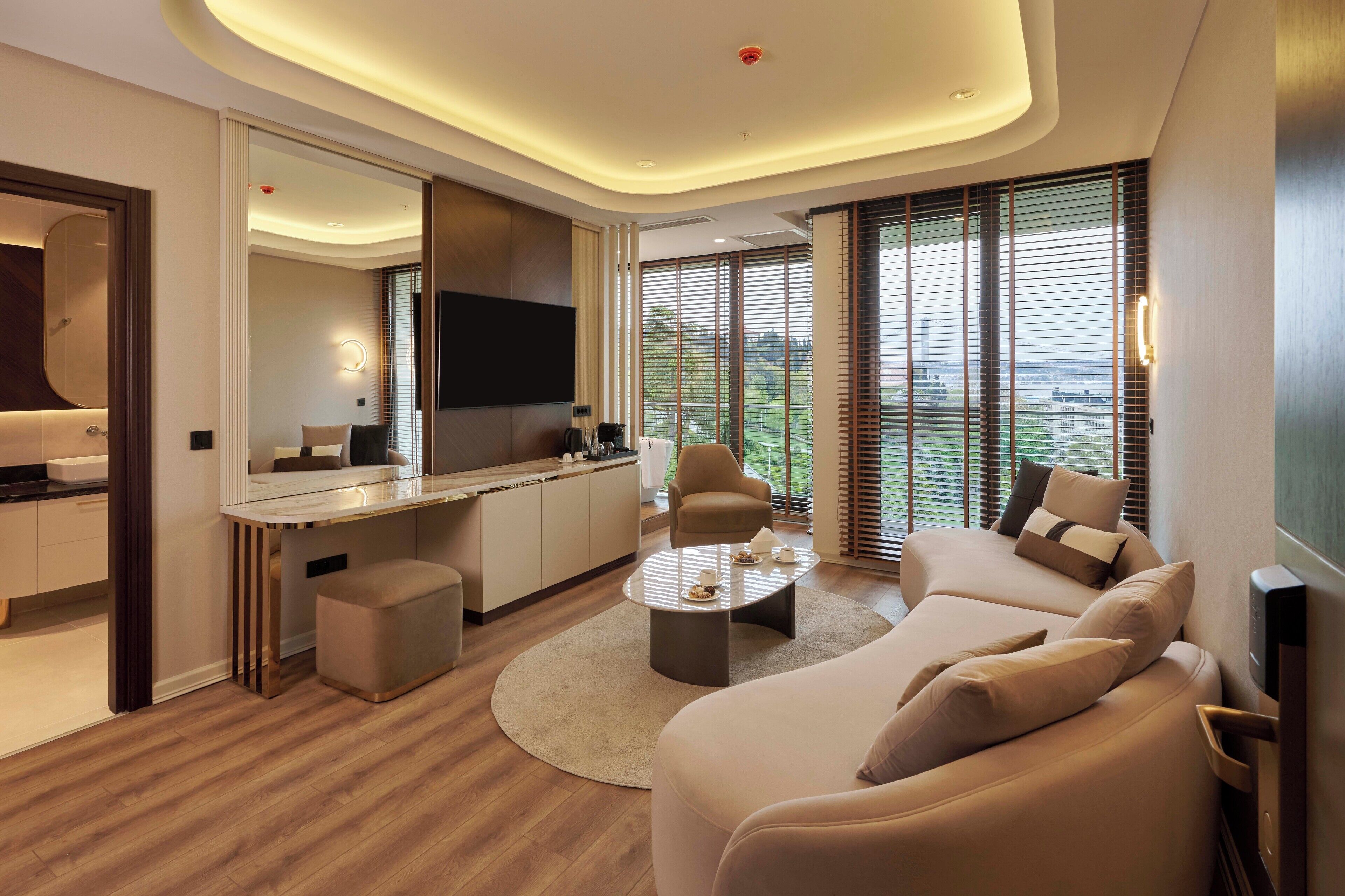 A modern living room with a sofa, coffee table, and a TV. There is a kitchen area with a dining table and chairs. The room has large windows with blinds.