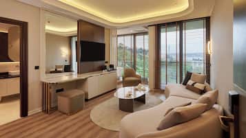 Ein modernes Wohnzimmer mit einer Couch, einem Couchtisch und einem Fernseher. Es gibt eine Küchenzeile mit einem Esstisch und Stühlen. Das Zimmer hat große Fenster mit Jalousien.