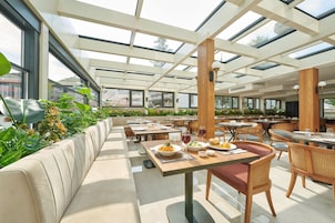 An outdoor dining area with multiple tables and chairs, surrounded by greenery and large windows.