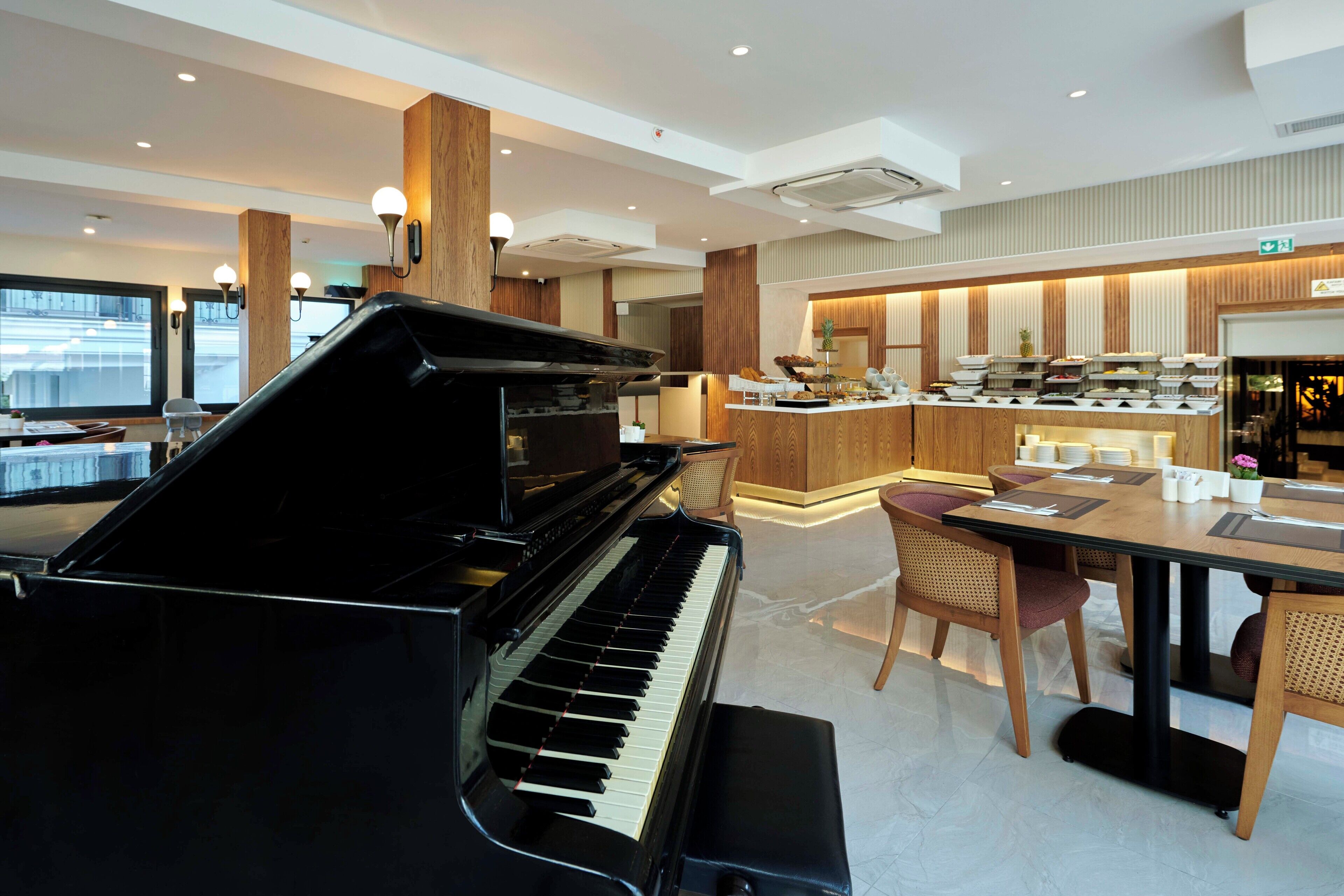 A modern hotel lobby with a grand piano, a buffet area, and a seating area with wooden chairs.