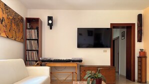 Een woonkamer met een witte bank, een houten salontafel met een plant, een boekenkast, een flatscreen televisie en een toetsenbord op een bureau.