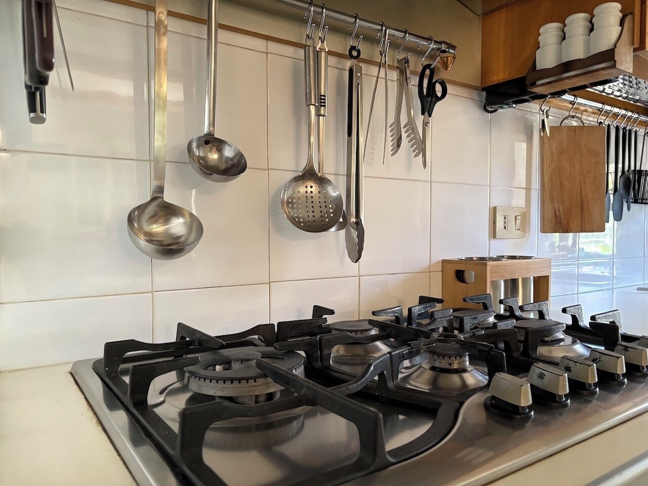 A kitchen with a gas stove, hanging utensils, and a wooden shelf with kitchenware.