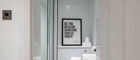 A modern bathroom with a glass shower enclosure, a toilet, and a sink. There is a framed quote on the wall.