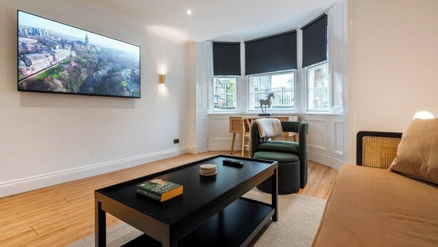 A modern living room with a large flat-screen TV mounted on the wall, a black coffee table with a book, a green armchair, and a beige sofa.