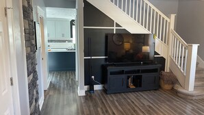 A modern living room with a staircase, a flat-screen TV, and a kitchen in the background. - Charming 3-bedroom, 2-12/ bath house with AC, WiFi in peaceful Fortville (Fortville)