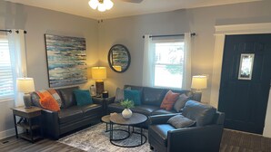 A living room with a dark leather sofa, a round coffee table, and two side tables with lamps. There is a painting on the wall, a window with curtains, and a dark door. - Charming 3-bedroom, 2-12/ bath house with AC, WiFi in peaceful Fortville (Fortville)