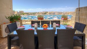 An outdoor dining area with wicker chairs and a glass table, overlooking a marina with boats and buildings. - Charming 1-bedroom house with AC, WiFi in beautiful Birzebbugia (Birzebbugia)