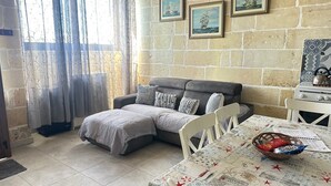 A cozy living room with a sofa, a dining table with a map-themed tablecloth, and a kitchen area with a stove. - Charming 1-bedroom house with AC, WiFi in beautiful Birzebbugia (Birzebbugia)