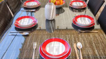 A glass-top dining table with red and white plates, black wicker chairs, and a fruit bowl in the center.