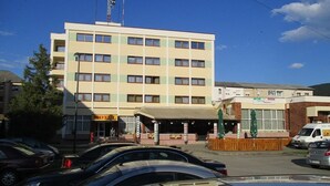 Ein mehrstöckiges Hotel mit schlichter Fassade, einer überdachten Eingangsparte und einem Parkplatz mit mehreren Autos.
