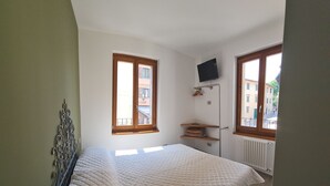 A bedroom with a bed, two windows, a radiator, and a TV on the wall.