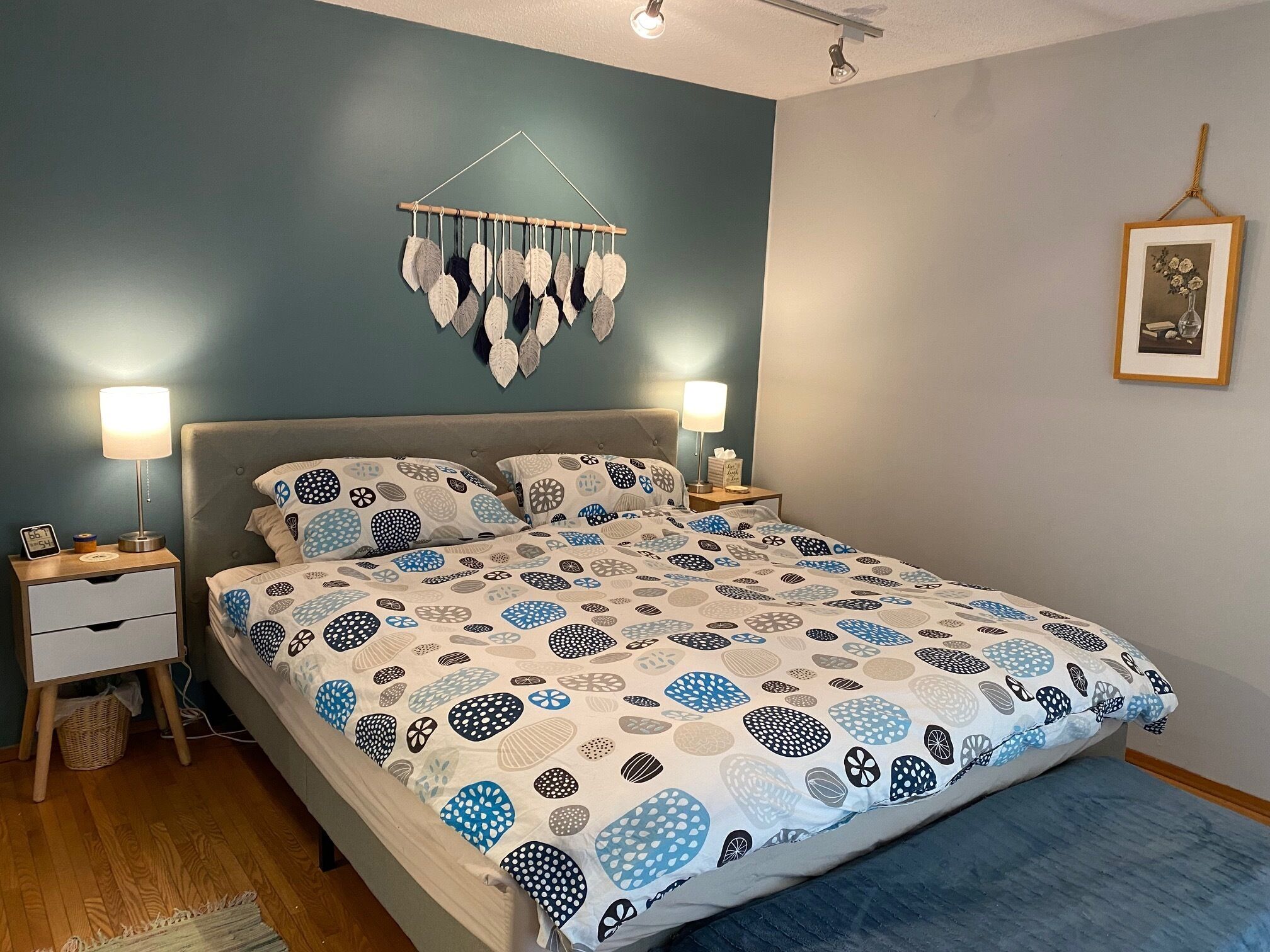 Un lit bien fait, avec une literie à motifs, deux tables de chevet avec des lampes, une décoration murale suspendue et un tableau encadré accroché au mur.