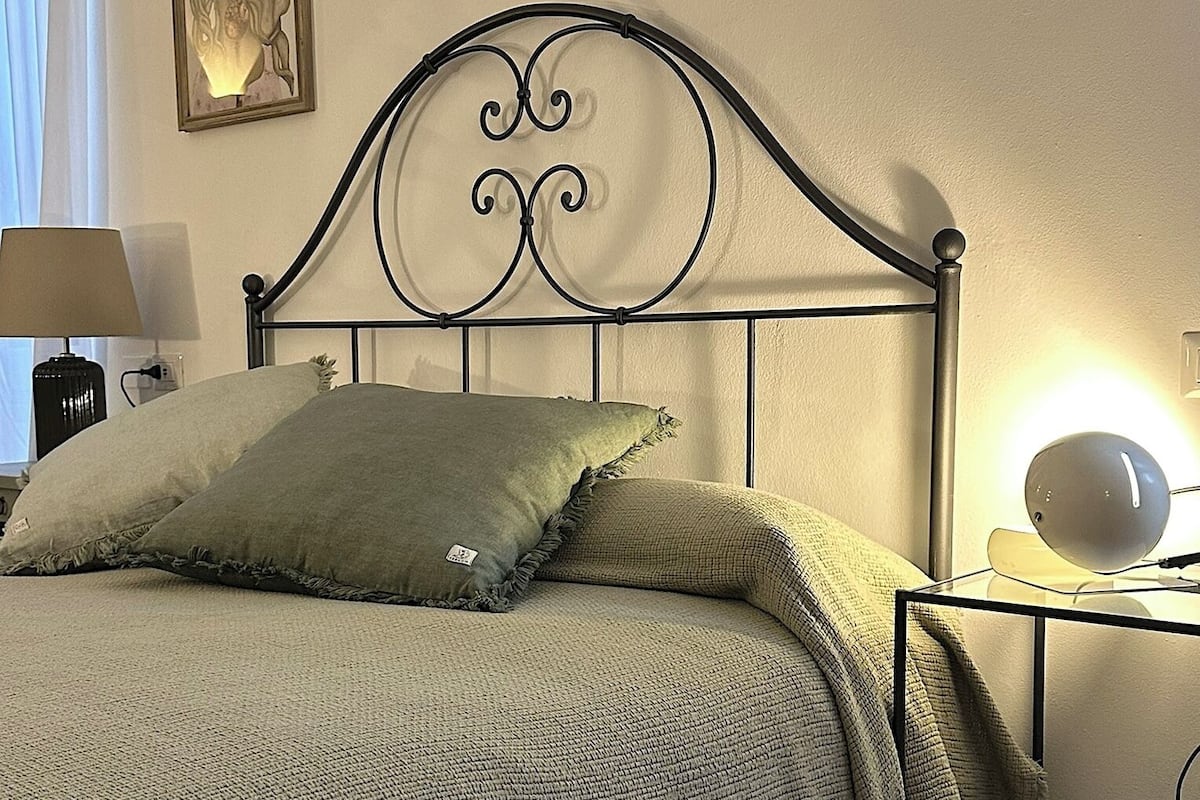 Een opgemaakt bed met een metalen hoofdeinde, een bedlampje en een wandlamp.