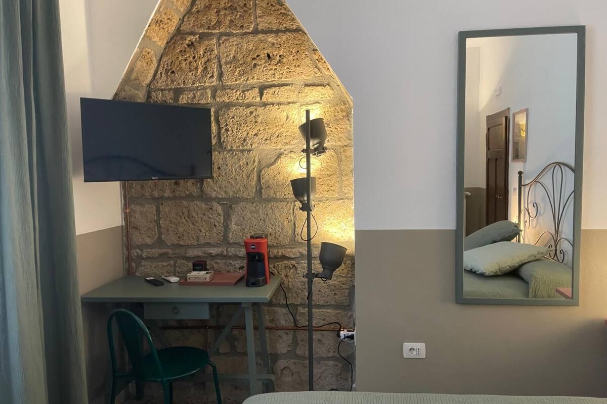 Een kleine kamer met een stenen muur, een bureau met stoel, een bed, een aan de muur bevestigde televisie en een spiegel die het bed reflecteert.