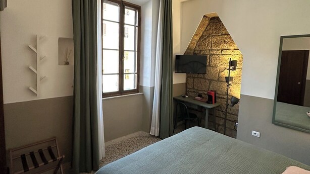 A bedroom with a bed, a desk, a chair, a window with curtains, a stone wall, and a mirror. - L'Altro Sapore Rooms (Pitigliano)