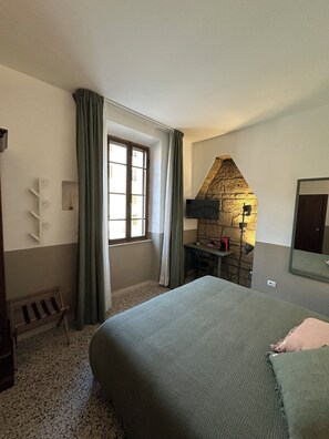 Une chambre à coucher comprenant un lit, un bureau, une chaise, une fenêtre avec des rideaux, un mur en pierre et un miroir.