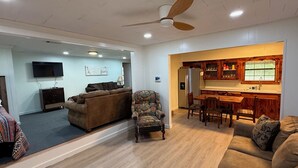 Amplia sala de estar con un sofá marrón, un sillón estampado y una mesa de comedor de madera con sillas. Se observa un ventilador de techo, y en el fondo, una cocina con armarios de madera y un refrigerador.